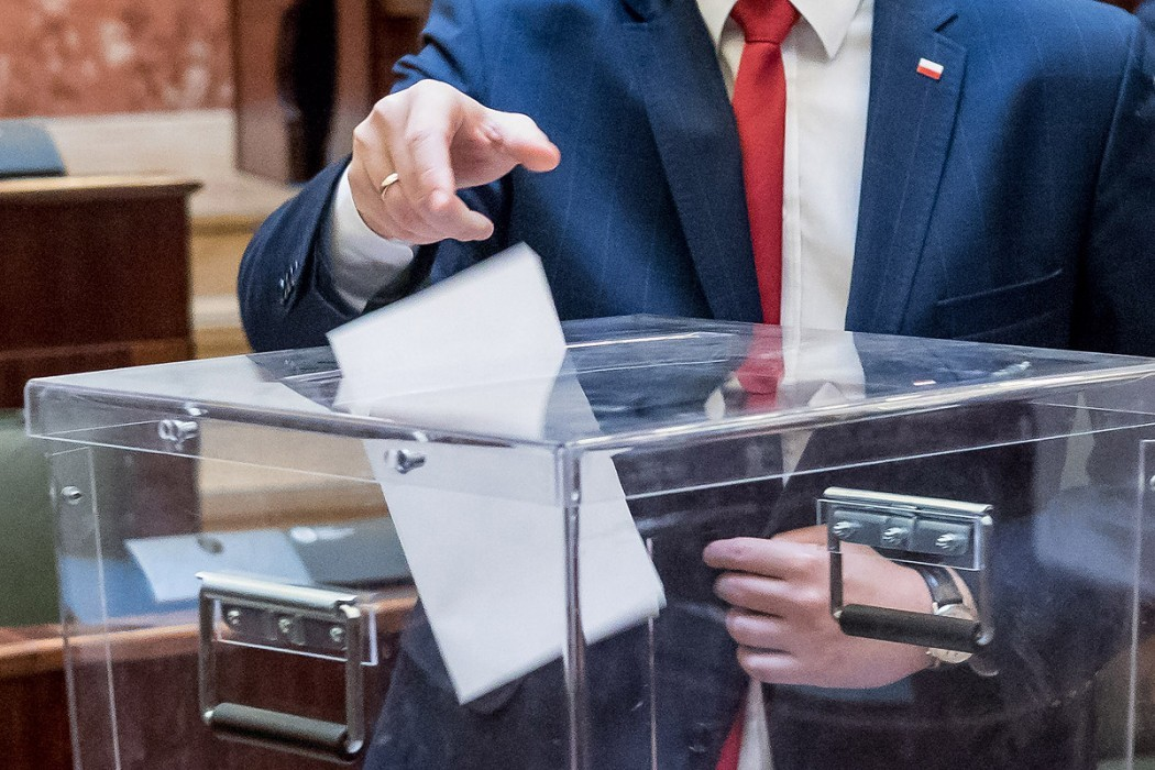 Zdjęcie przedstawia osobę wrzucającą kartę wyborczą do urny